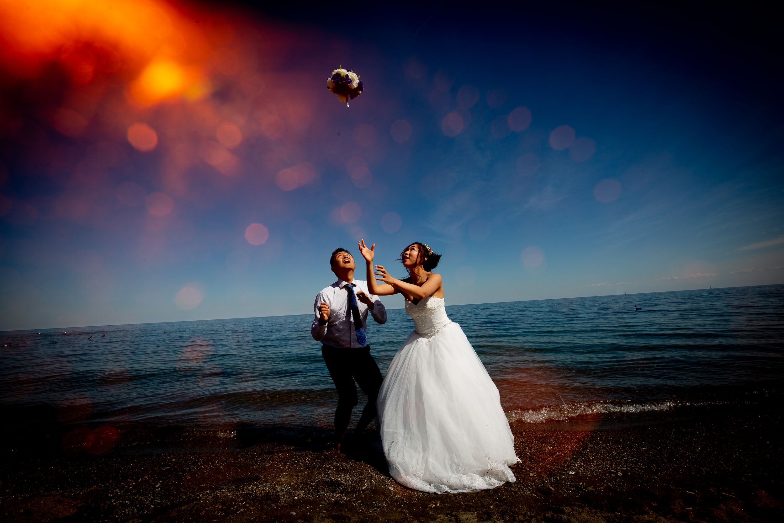 Dramatic sunset wedding photo at beach with couple tossing bouquet against orange sky.