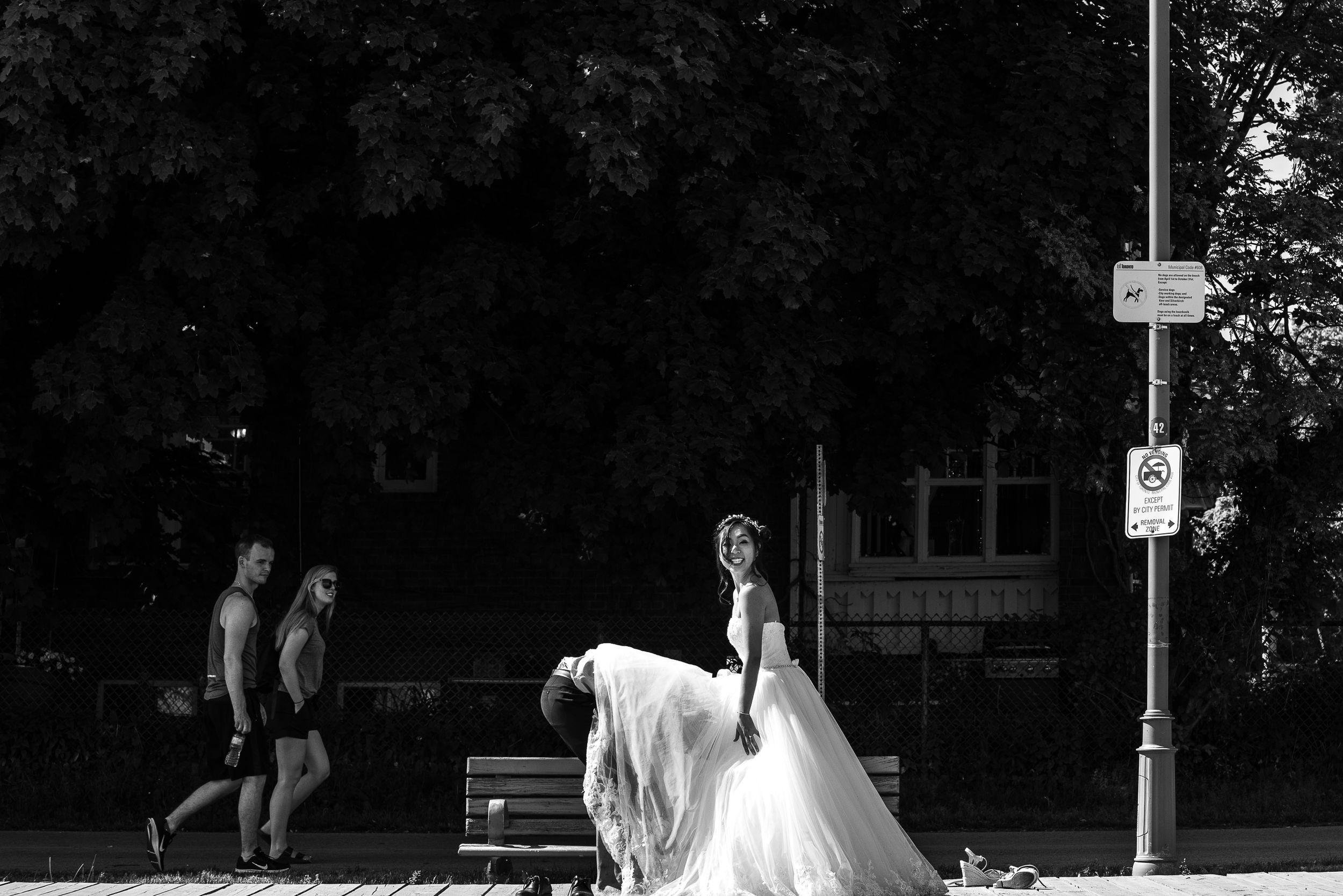Black and white photo of a bride in a flowing white dress walking past a park bench.