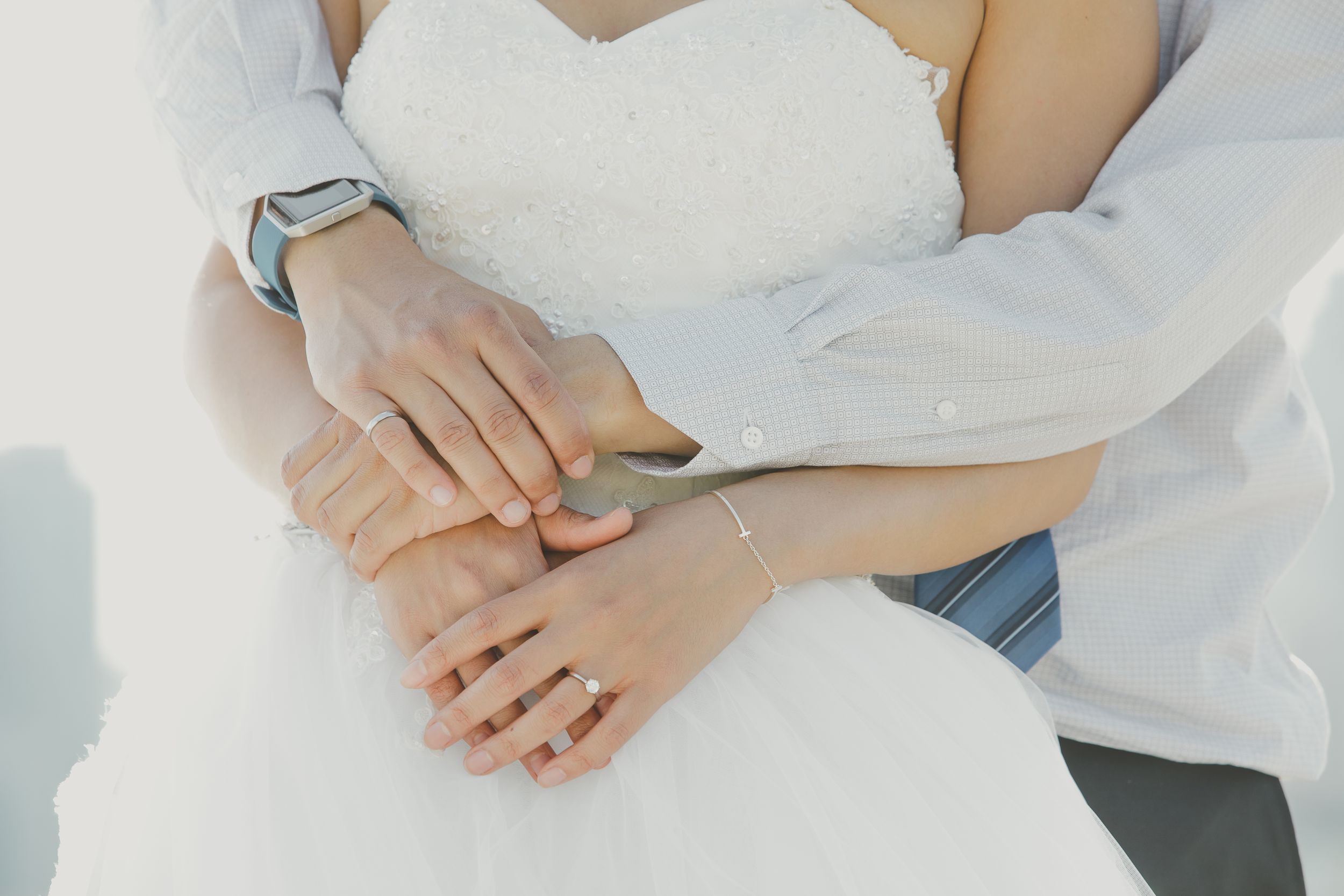 Close-up detail of hands with wedding rings resting on white dress fabric.