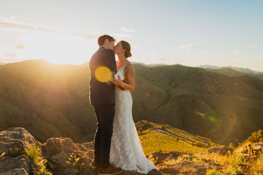 A couple in wedding attire embraces on a mountaintop during golden hour as the sun sets over rolling hills.