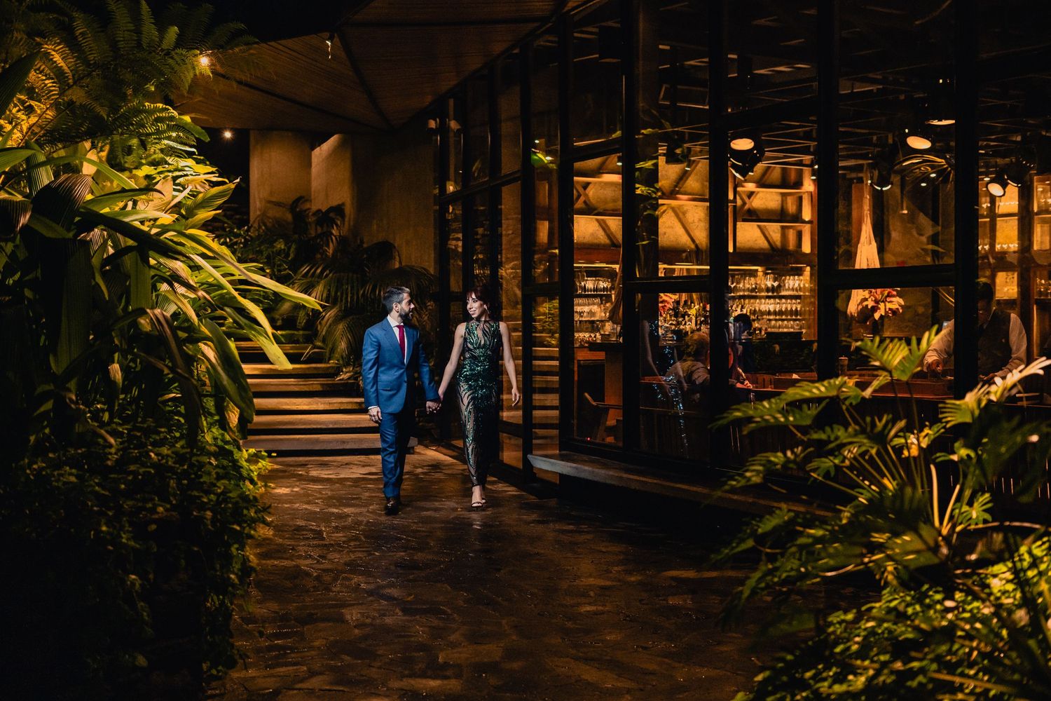 A couple walks along an illuminated garden path with tropical plants and glowing architectural features at night.