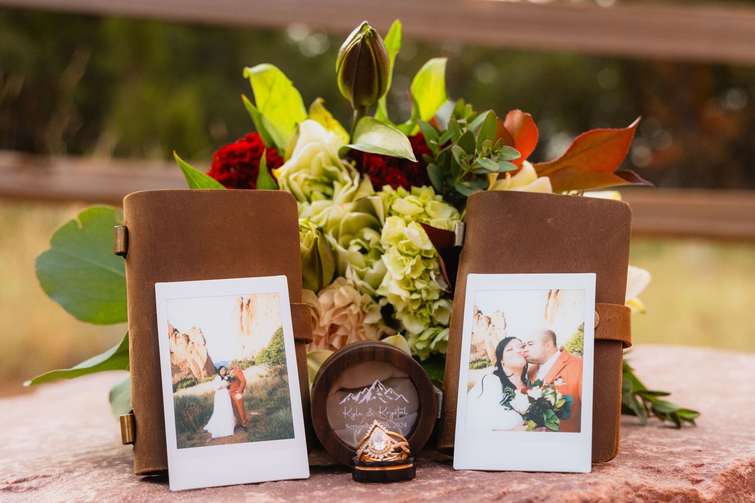 A brown frame and vase centerpiece with instant photos and fresh yellow and red flowers on a pink surface.