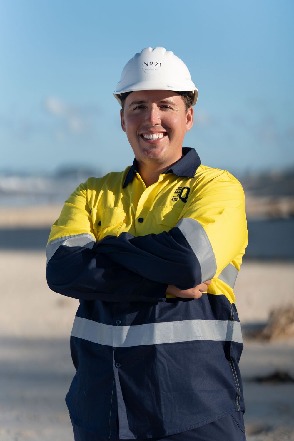 Worker in high-visibility yellow and navy uniform with white hard hat standing outdoors with arms crossed.