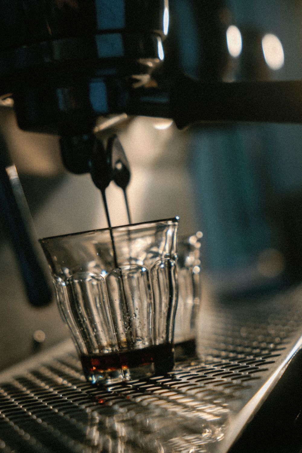 Espresso machine pours dark coffee into a ribbed glass cup on a metal grate with bokeh lights in background.