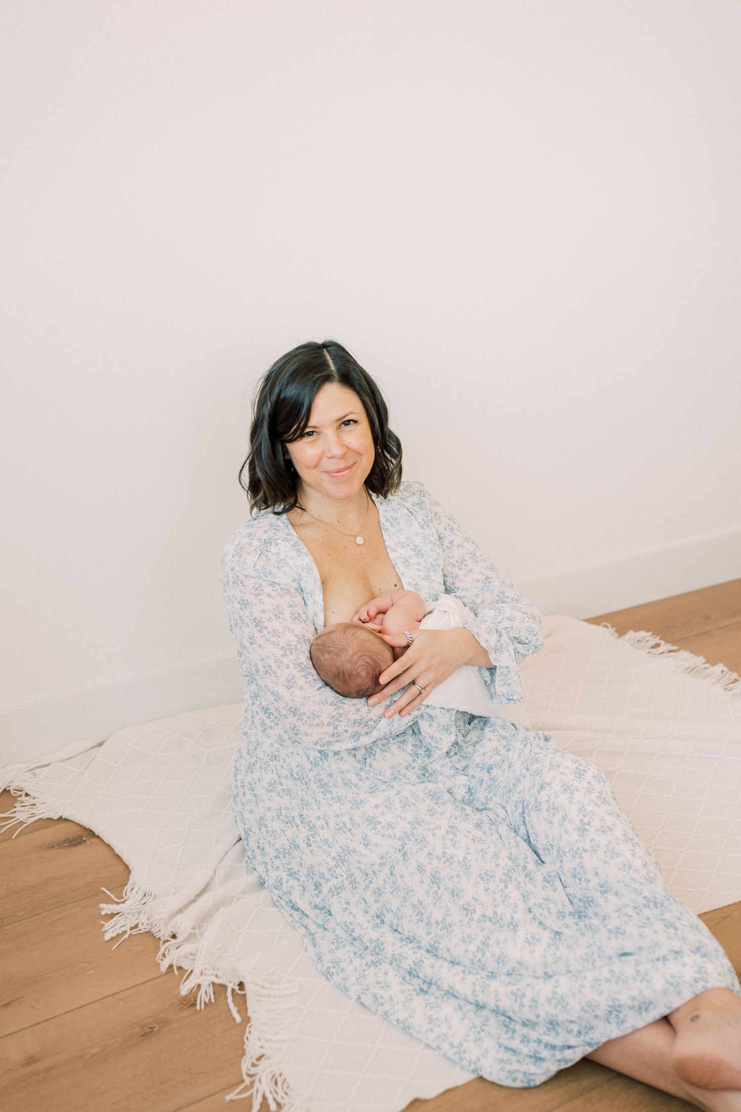 A mother wearing a light blue floral dress sits on wood flooring while nursing a baby.
