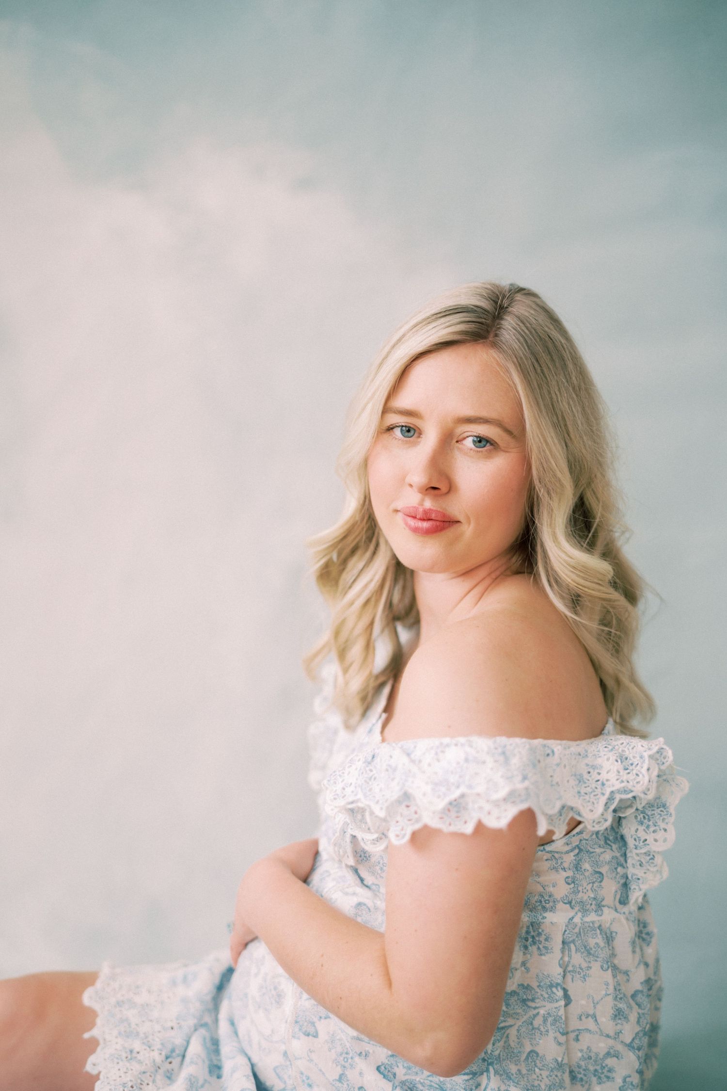 A soft and ethereal portrait featuring a blonde woman in an off-shoulder lace dress against a pastel backdrop.