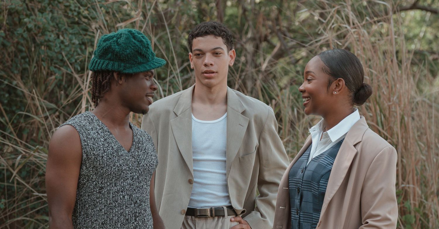Three people in casual and formal attire having a conversation outdoors against a natural background.