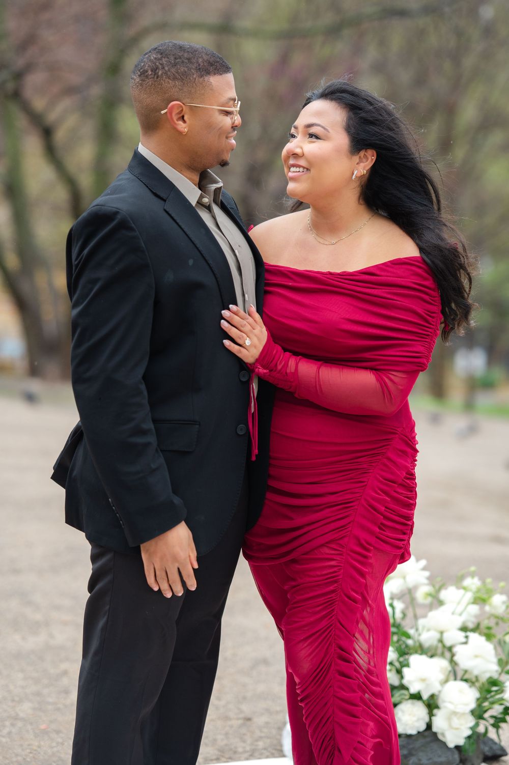 Romantic couple smiling at each other outdoors, with woman in red ruched gown and man in black suit after proposal moment at DUMBO, NYC