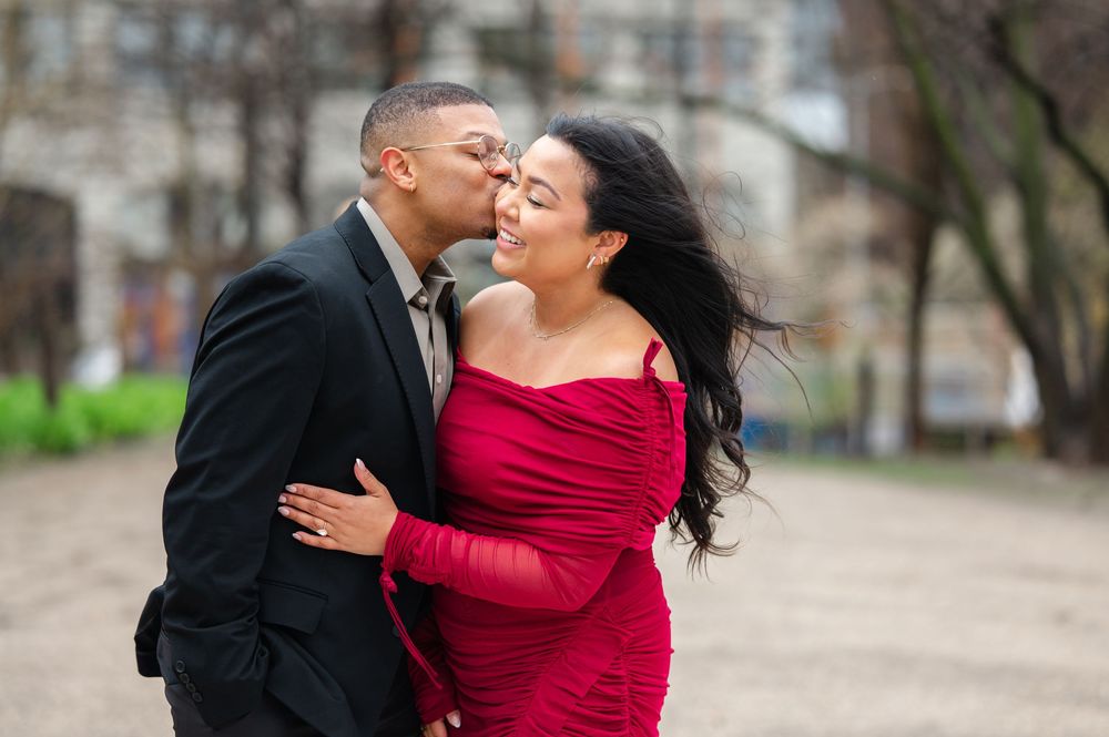 A couple shares a romantic moment outdoors, with the woman wearing a vibrant red dress and the man in a black suit in front of DUMBO