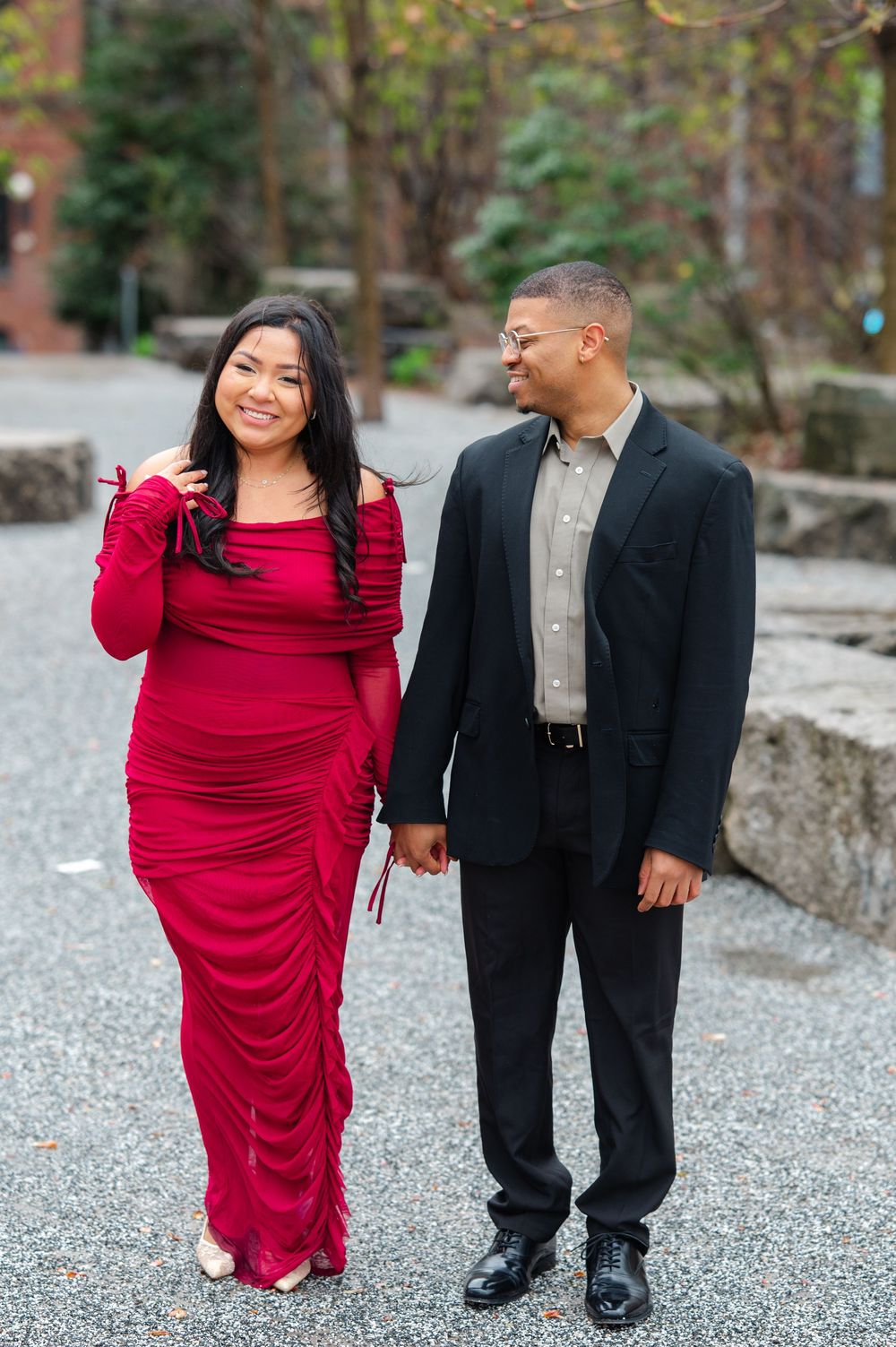 A couple in formal attire walks together on a gravel path, with the woman wearing a red dress and man in black suit after engagement