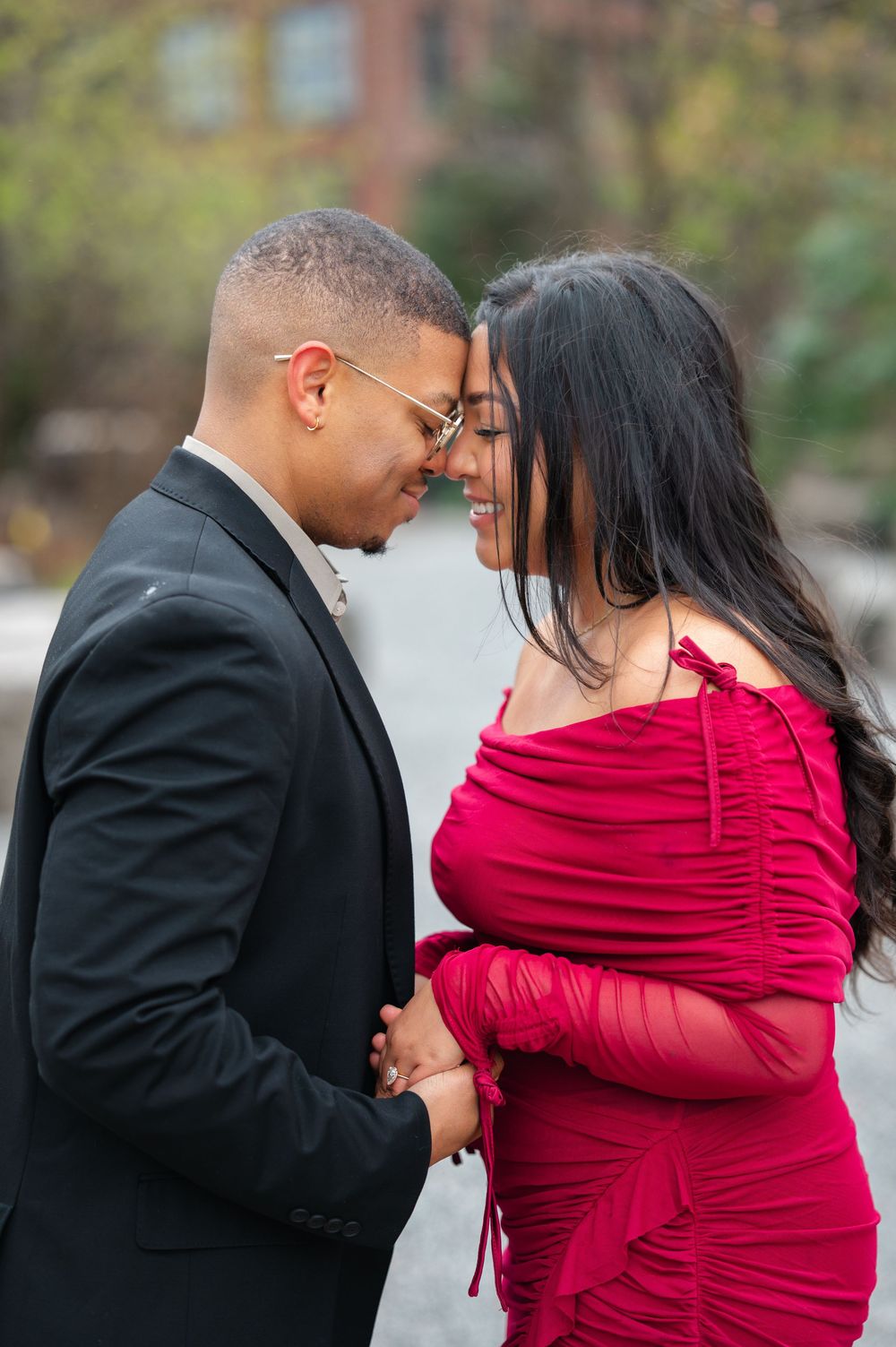 A couple in formal attire share an intimate moment outdoors, with the person in a red ruched dress and another in a black suit.