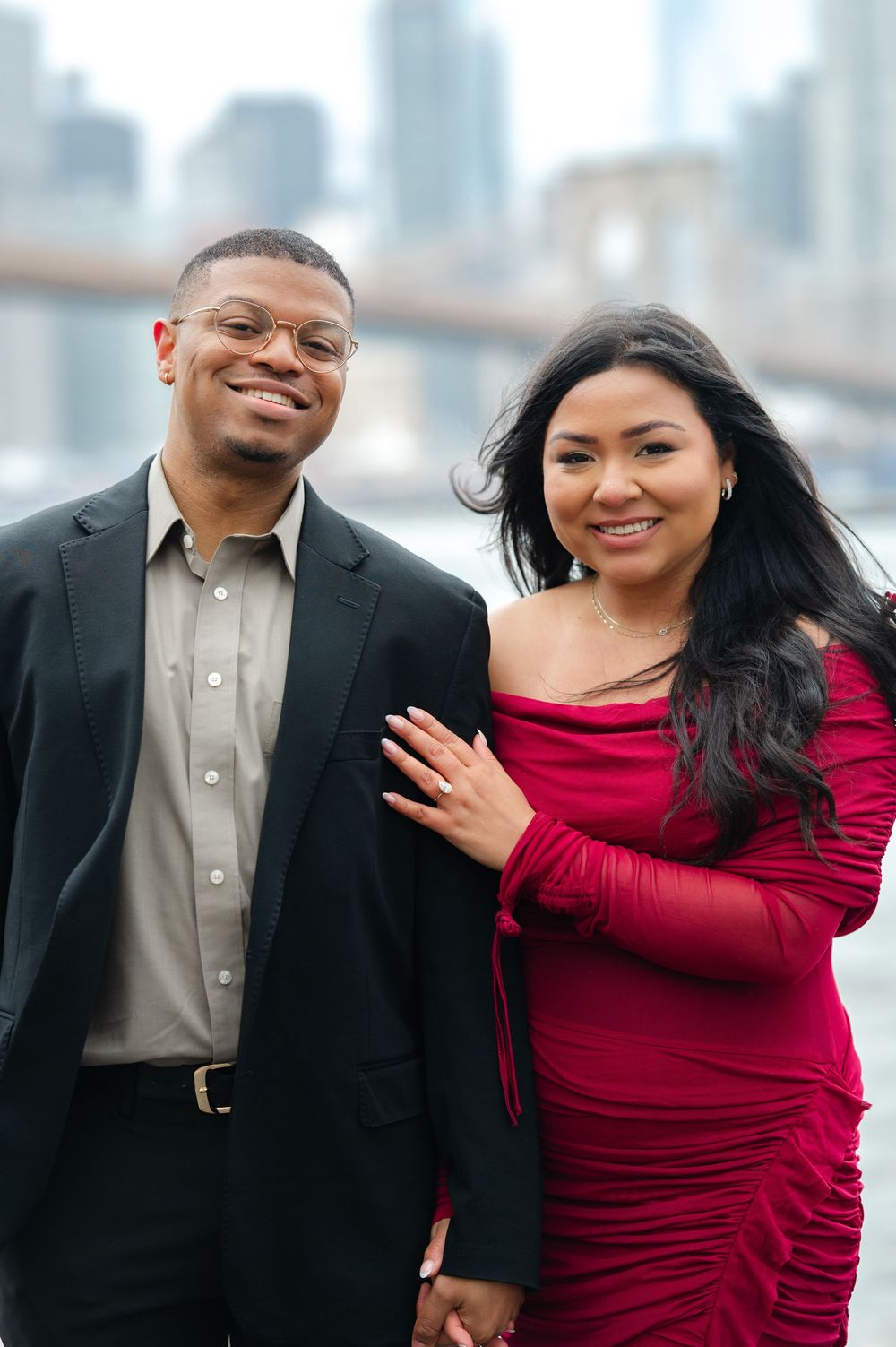 A couple poses together outdoors in formal attire with a city skyline in the background after proposal