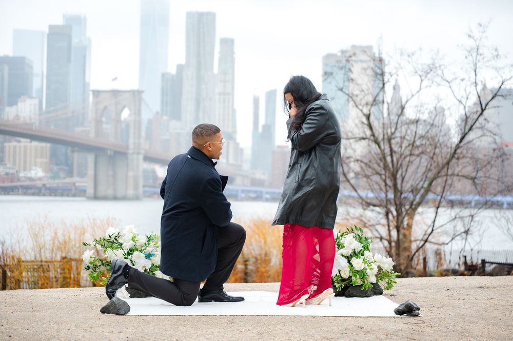 A couple shares a romantic proposal moment along the Brooklyn waterfront with the city skyline and bridge in the background.