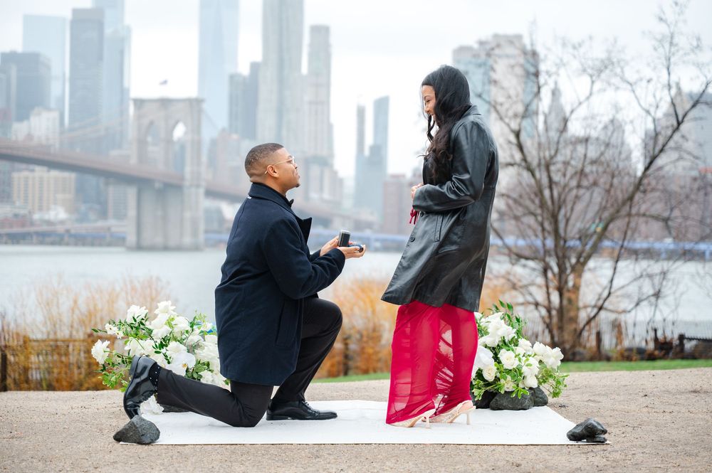A romantic proposal scene at a waterfront park in New York City with the Brooklyn Bridge and skyline in background at DUMBO.