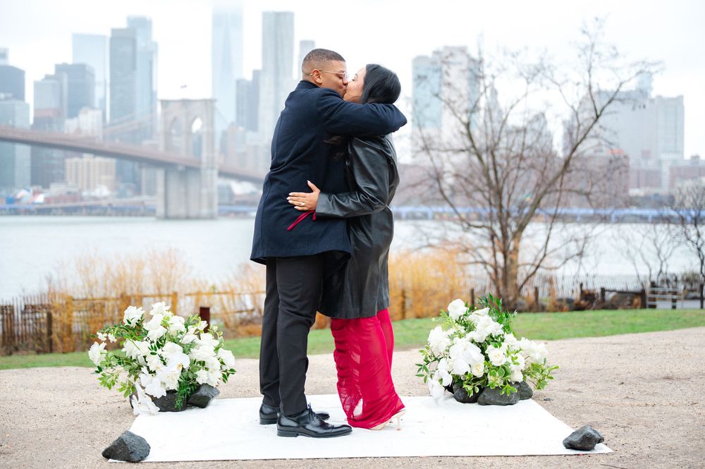 A couple embraces during their engagement moment by the waterfront with white floral arrangements and city views.