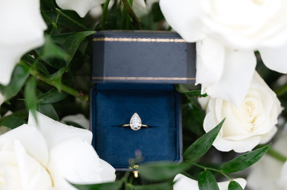 Pear-shaped diamond engagement ring in navy blue velvet box surrounded by white roses and green leaves.