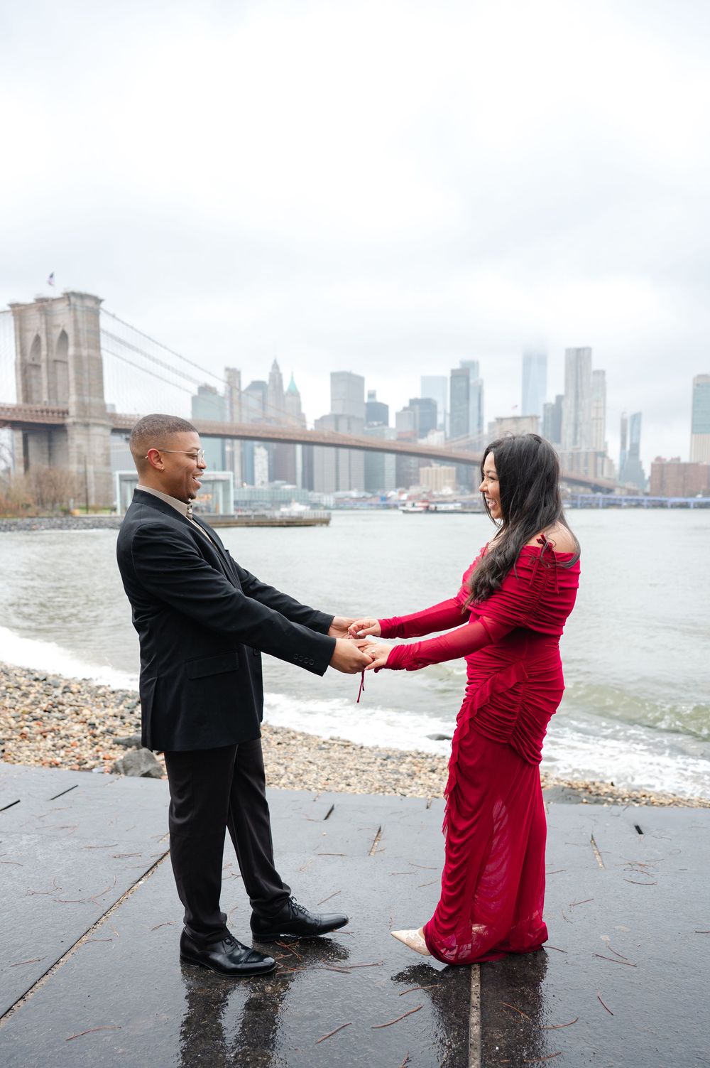 A couple in formal attire holds hands by the Brooklyn Bridge and Manhattan skyline on a cloudy day.