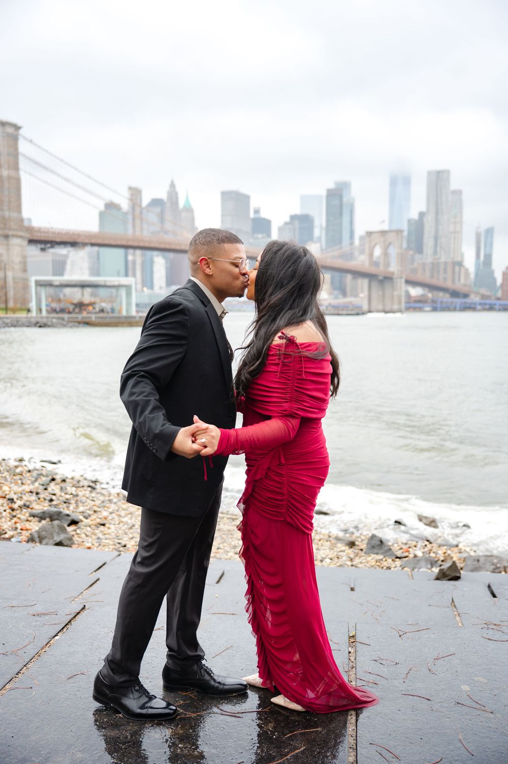 A couple in formal attire embracing by the Brooklyn Bridge waterfront with Manhattan skyline in background after proposal moment