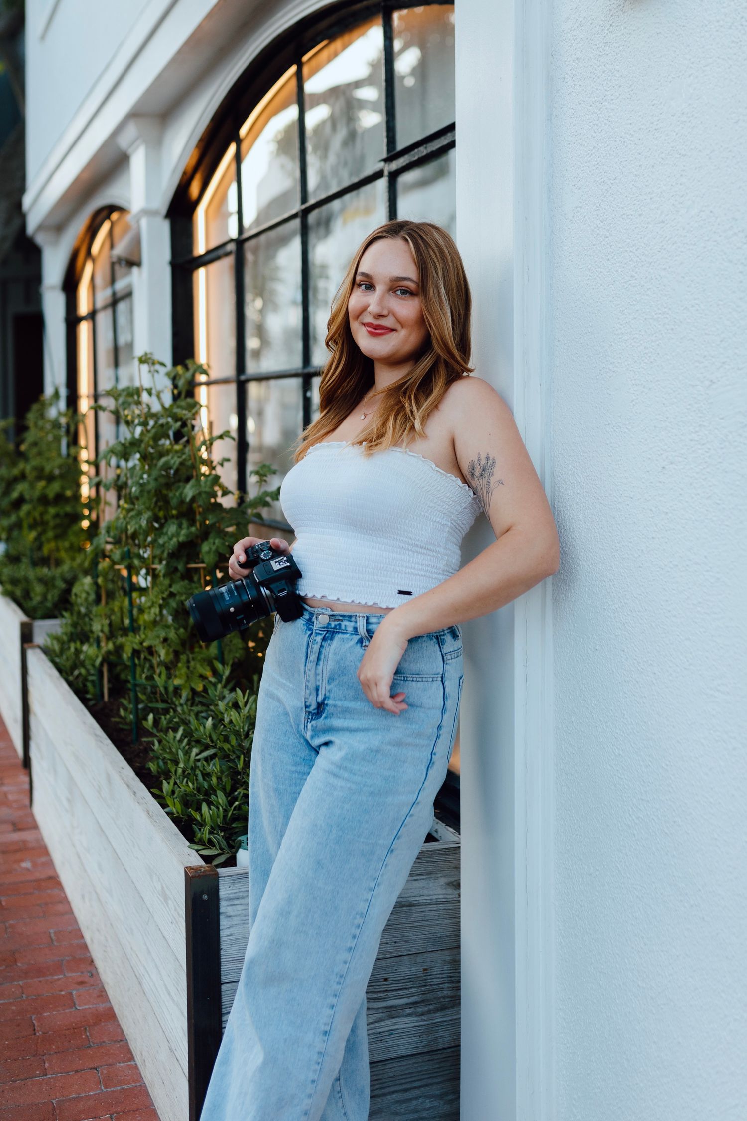 Photographer in casual white top and light wash jeans holds camera while standing by arched windows and greenery.