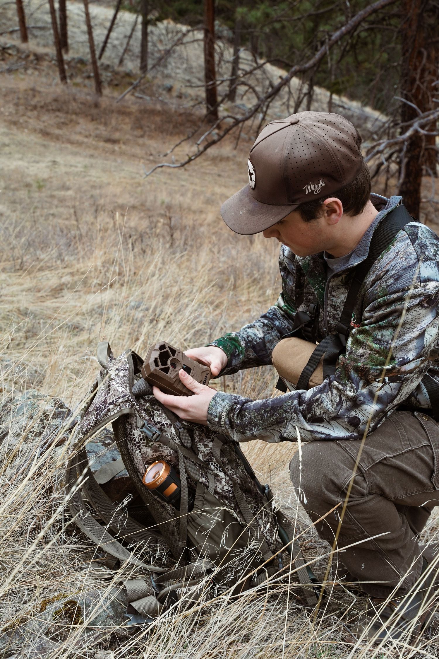 A hunter in camouflage clothing examines gear while kneeling on a hillside in dry grass and forest terrain.