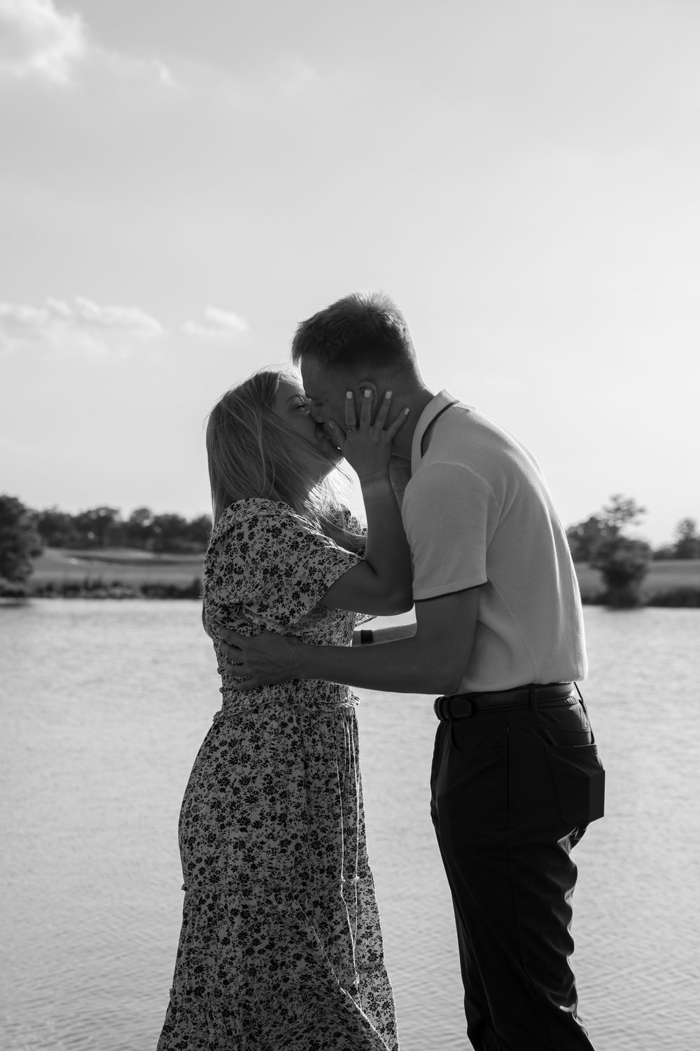 A black and white photograph of a couple embracing by a lake on a sunny afternoon.