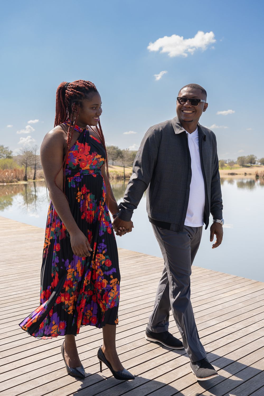 A couple walks hand in hand on a wooden boardwalk by a lake on a sunny day.