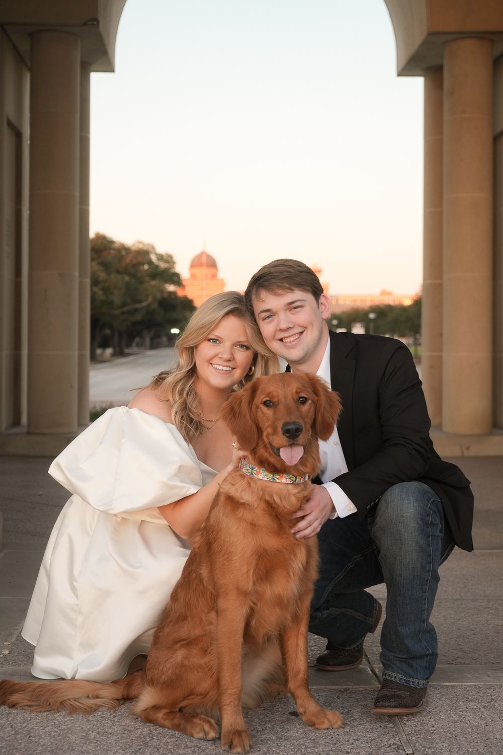 Couple poses with their golden retriever under an architectural arch at sunset.