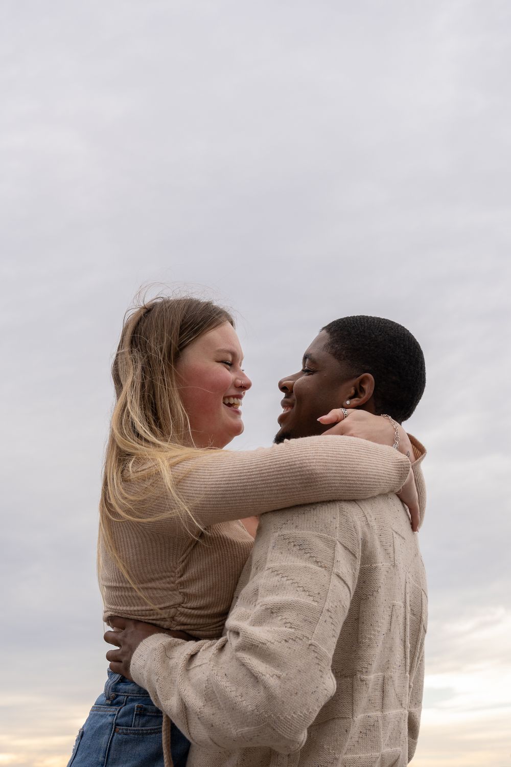 A couple embraces and laughs together against an overcast sky wearing neutral-colored sweaters.