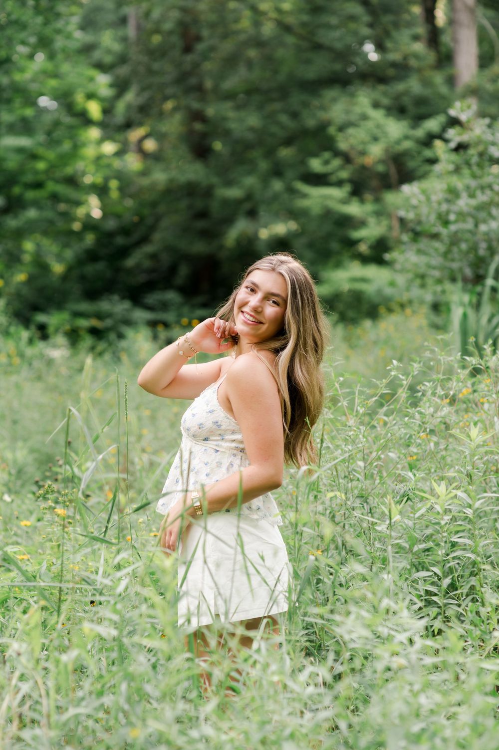 A person in a white summer dress poses playfully among tall grass and wildflowers in a sunlit woodland setting.