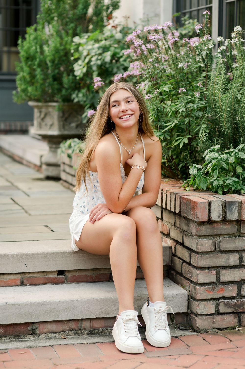 A person in white sneakers sits casually on brick steps surrounded by blooming summer flowers.