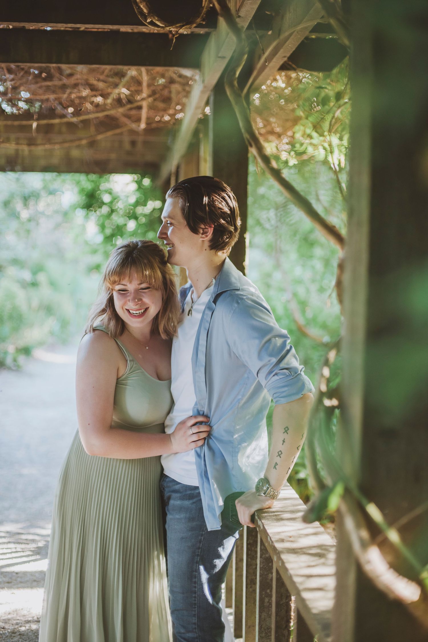 A couple laughs together, standing in a wooden arbor surrounded by greenery, with warm sunlight filtering through.