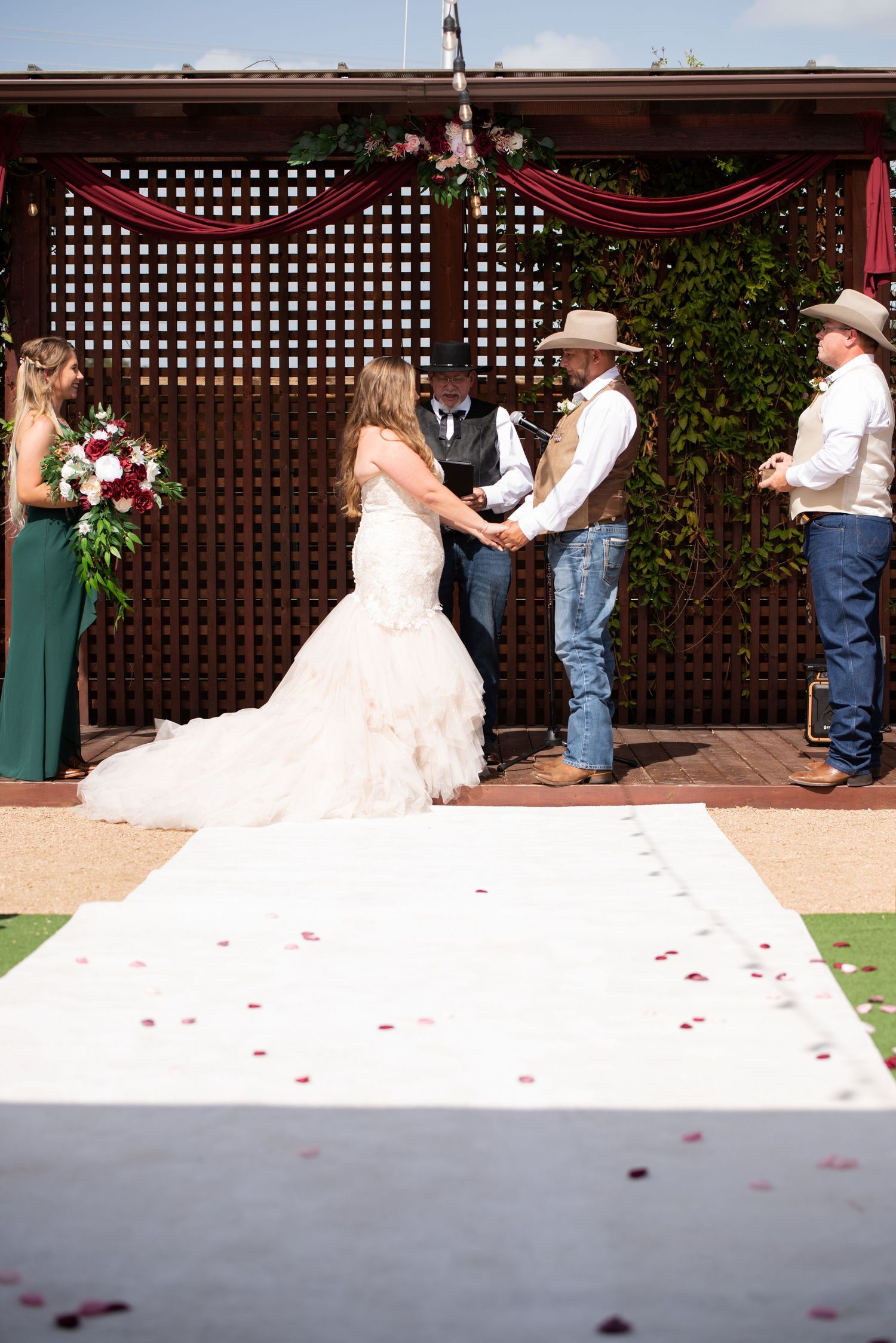 An outdoor wedding ceremony takes place under a wooden lattice pergola decorated with red fabric and rose petals on the aisle.