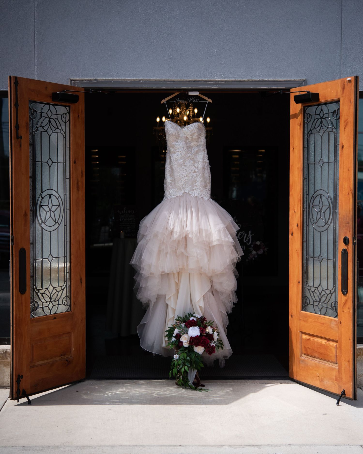 A white mermaid-style wedding dress hangs elegantly in a doorway with stained glass panels and a bouquet below.