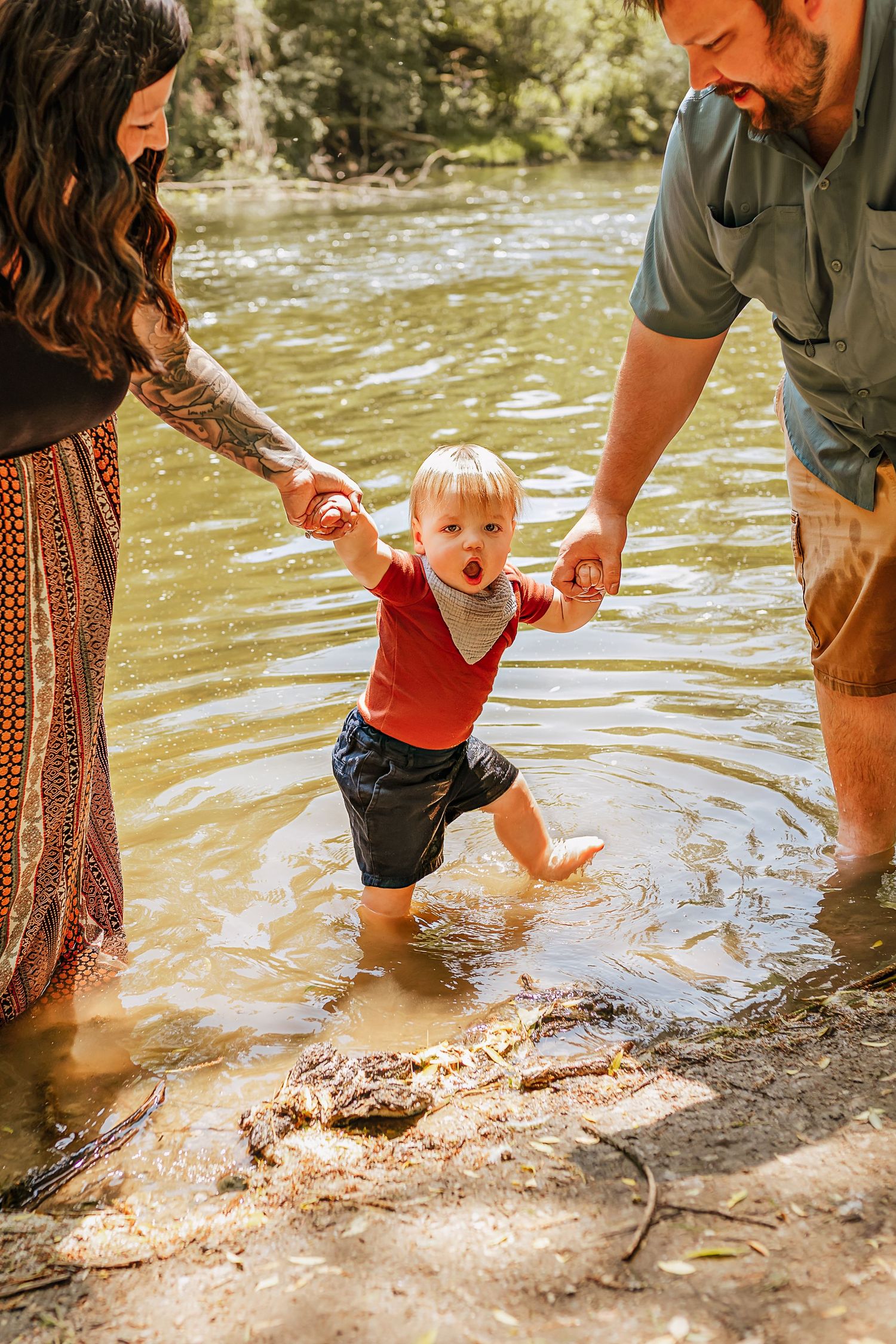 A toddler in a red shirt splashes in shallow river water while being supported by adults on a sunny summer day.