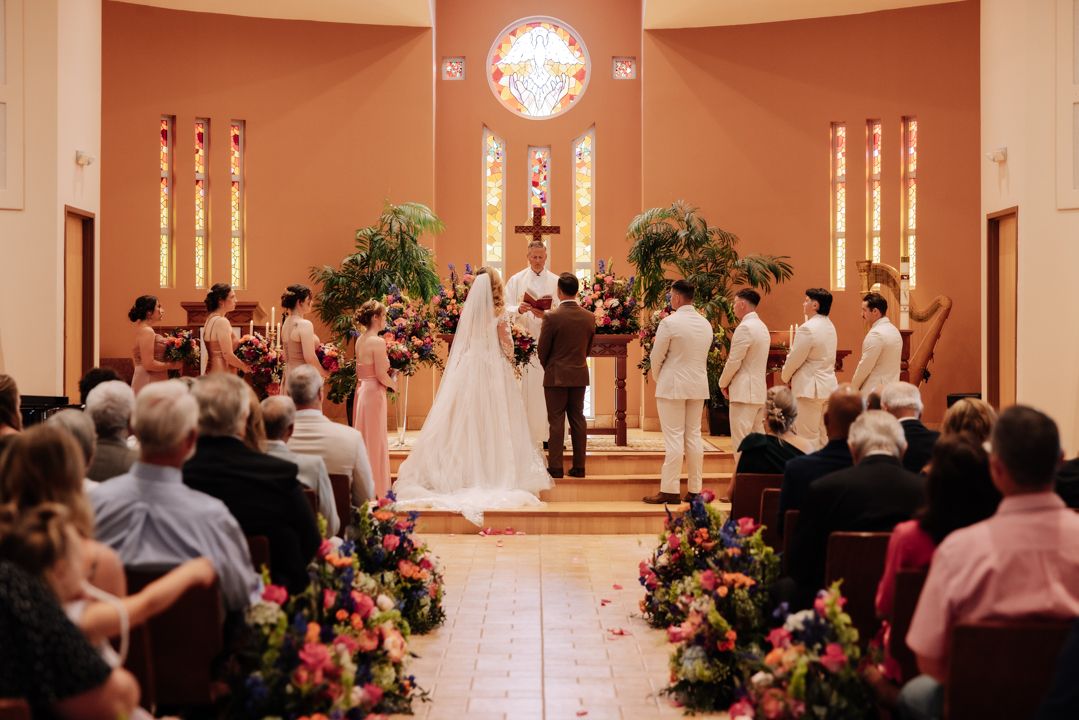 A wedding ceremony takes place in a church sanctuary with decorated floral arrangements and lit candles at the altar.