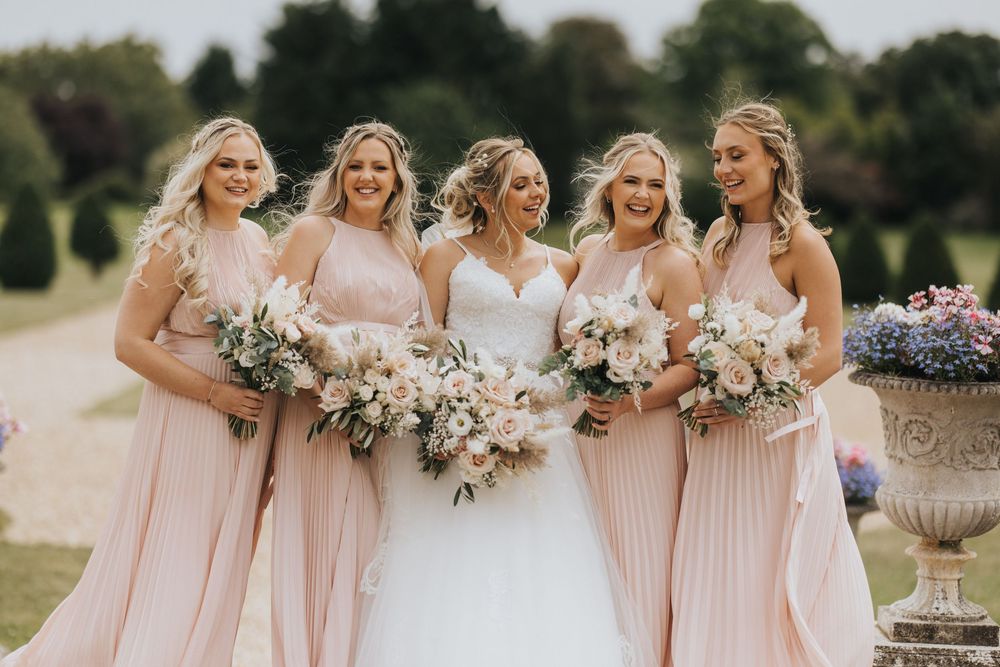 A bridal party in blush pink bridesmaid dresses and white wedding gown hold floral bouquets outdoors on sunny day.
