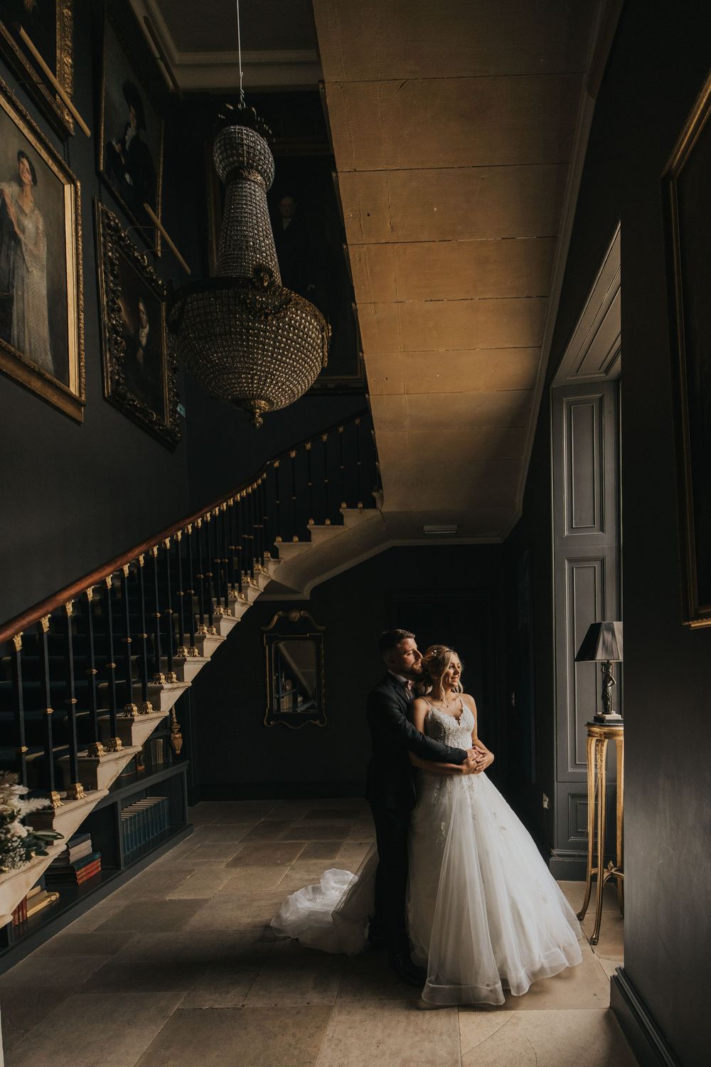 A romantic wedding photo in a dark hallway with an elegant chandelier and sweeping staircase.