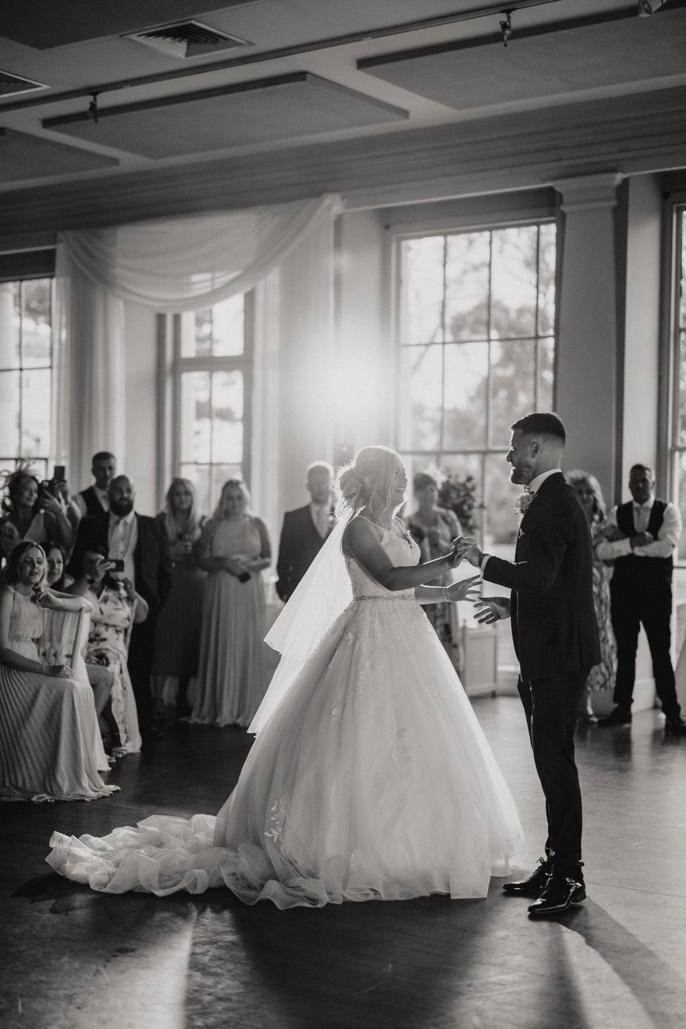 A black and white photo of a couple's first dance in a bright room with large windows.