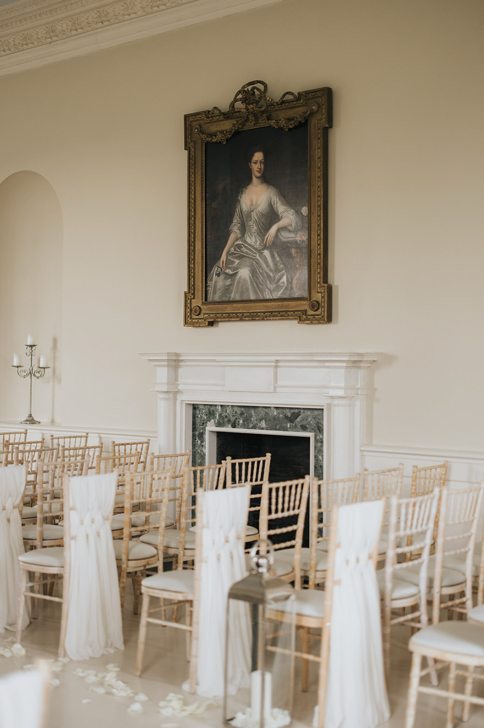 Empty white chiavari chairs arranged in rows inside an elegant room with antique portraits on the walls.
