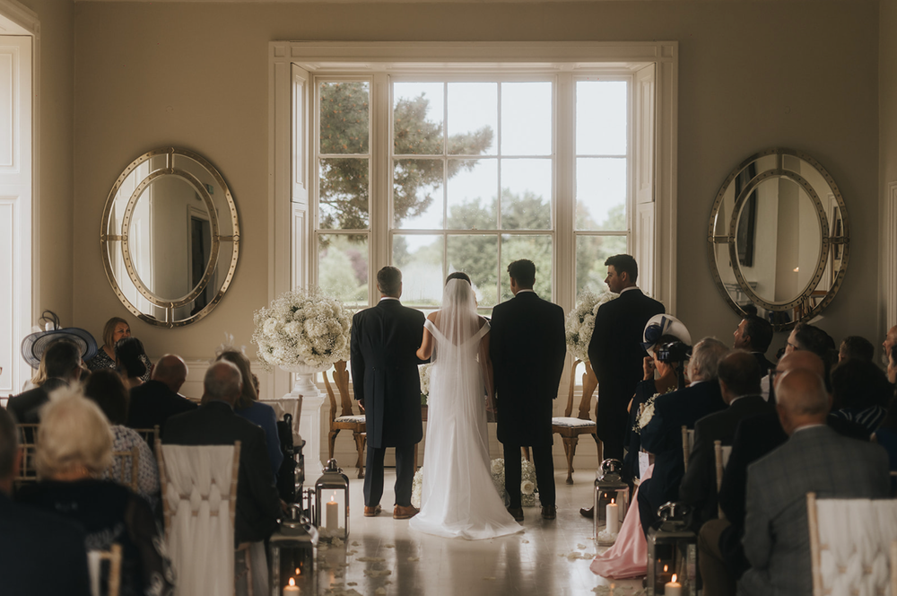 Wedding guests gather in an elegant sunlit venue with round mirrors and large windows for a special ceremony.