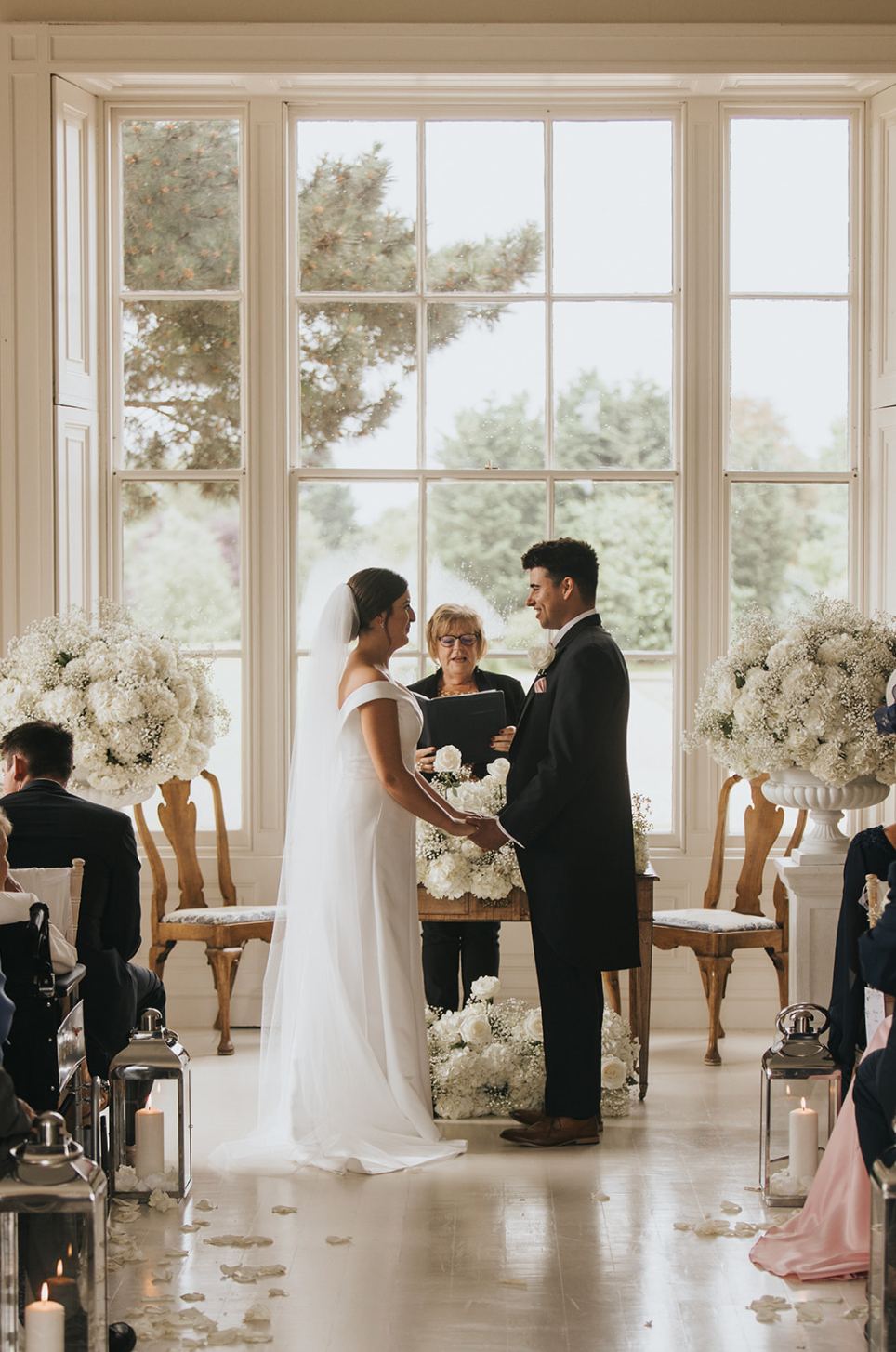 Wedding ceremony taking place in an elegant bright room with large windows and white floral decorations.
