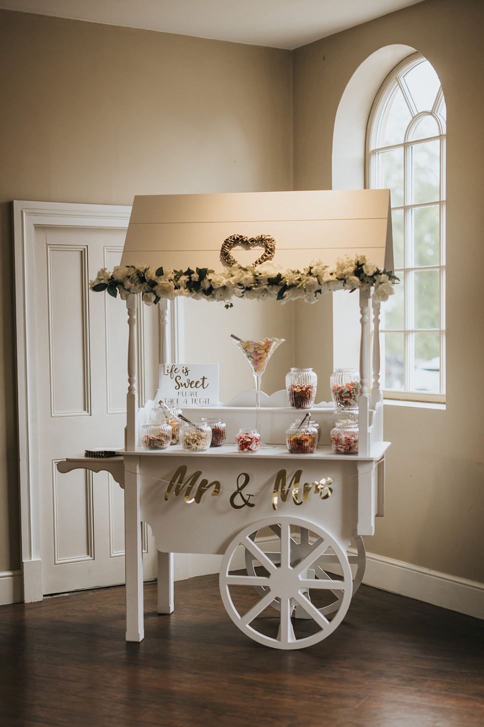 Elegant white dessert cart decorated with floral garlands and Mr & Mrs signage for a wedding reception.