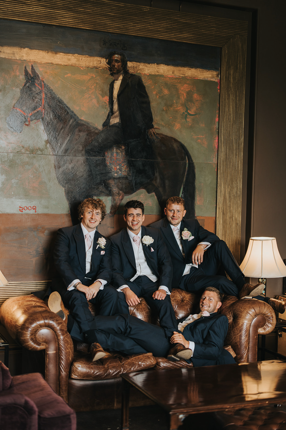 Four men in tuxedos lounging on brown leather chairs during a formal event.