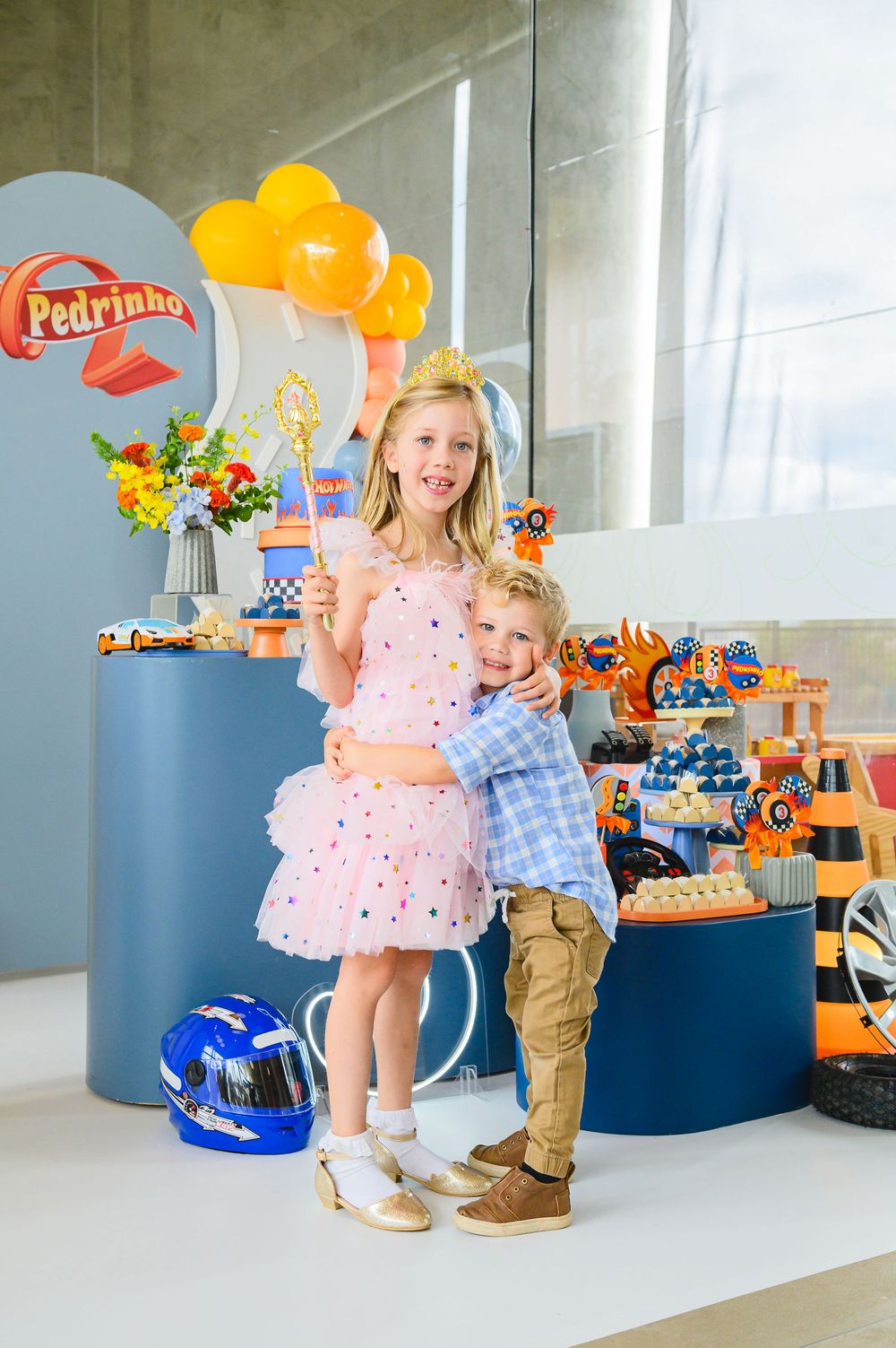 Children stand together at a birthday party with blue tables, orange balloons and toy decorations in the background.