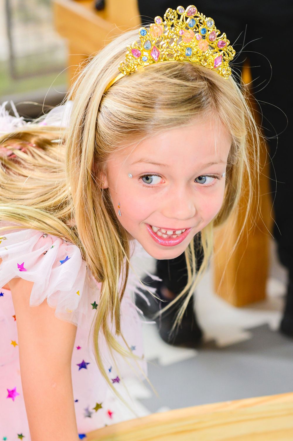 Young child wearing sparkly princess crown and star-patterned dress smiling brightly at camera.