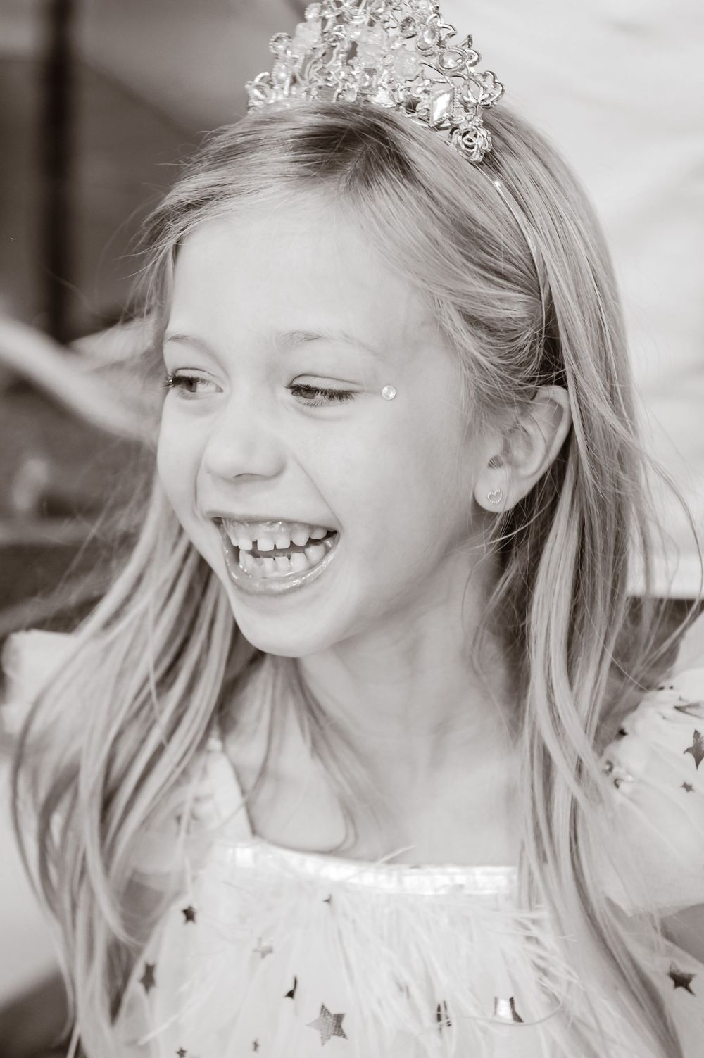 Black and white photo of child wearing tiara and laughing joyfully while wearing star-decorated dress.
