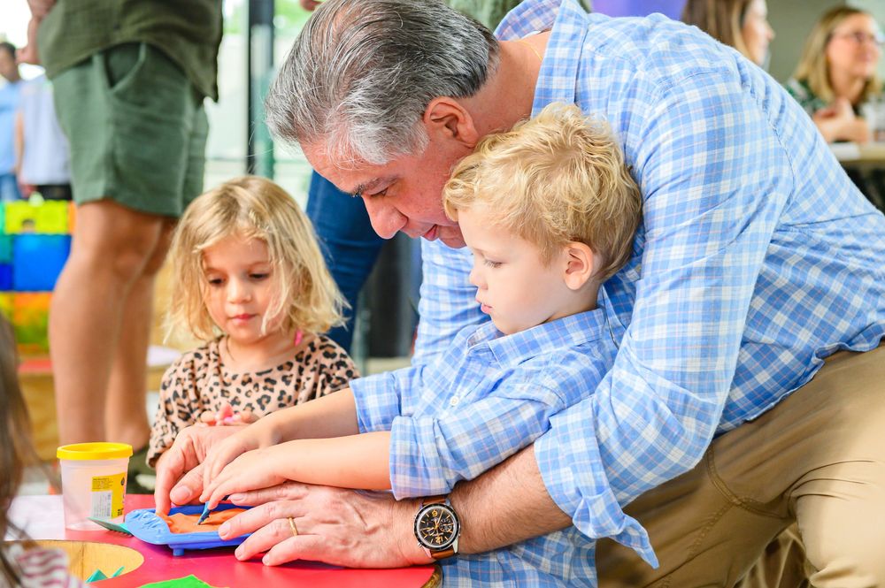 An older gentleman assists young children with a craft activity at a table.