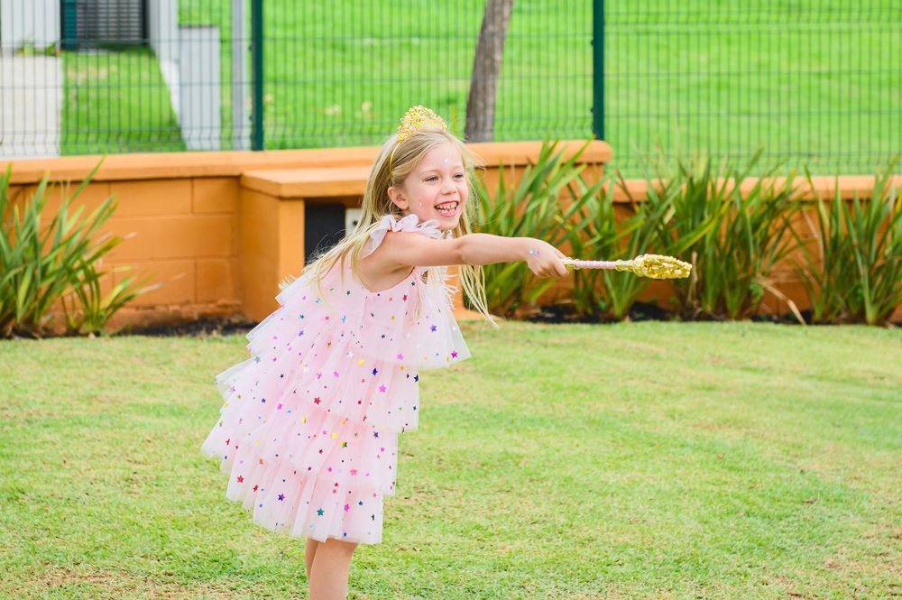Child in a pink polka dot dress dances playfully on green grass in a garden setting.