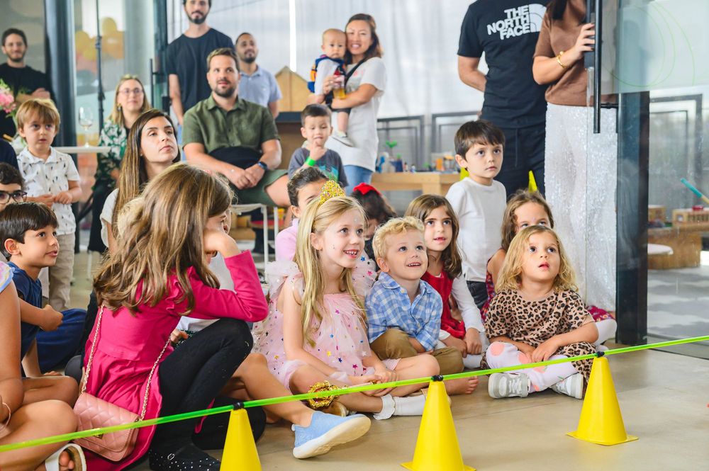 Children sit on the floor watching attentively during an indoor activity with yellow cones and adults in background.