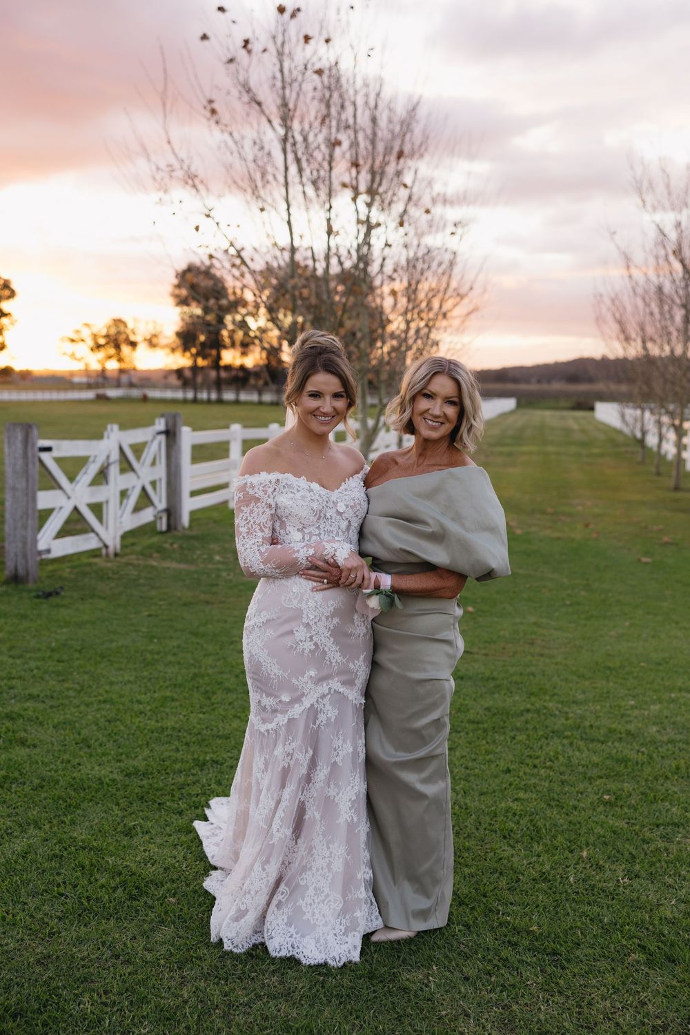 A sunset wedding portrait by a white fence featuring an off-shoulder lace gown and elegant evening attire.